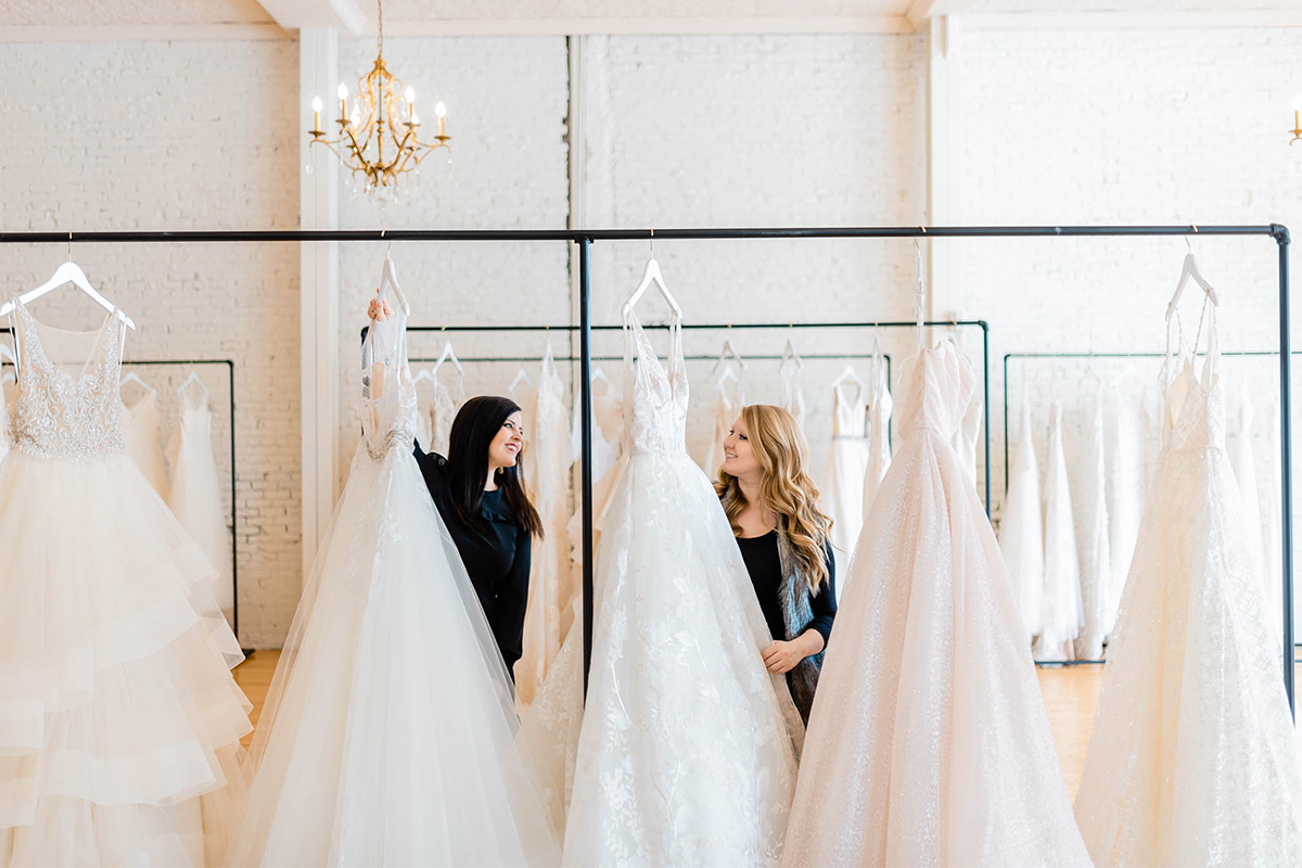 Kayla Thompson and Erin Rallis standing behind a rack of wedding dresses.