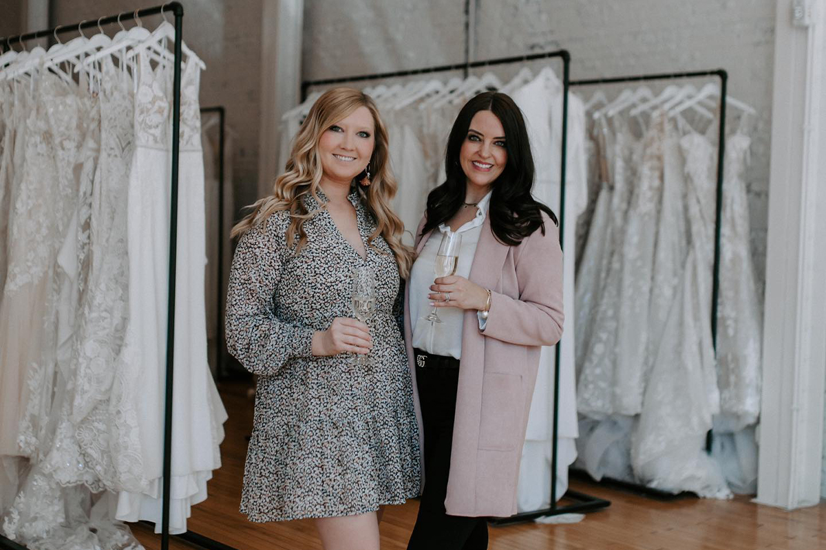 Erin Rallis and Kayla Thompson holding glasses of champagne in their small business, Marie and Marie Bridal.