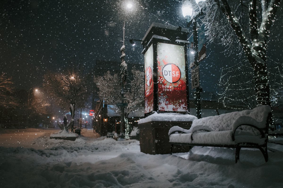 A bench covered in snow next to a lit up downtown Sioux Falls sign, with Christmas lights and falling snow in the background.