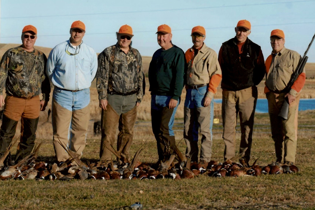 Seven men dressed in hunting gear, holding guns, and standing behind a row of dead pheasants.