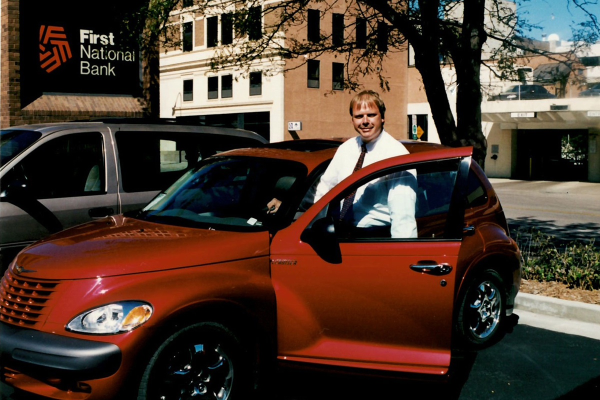 A young Russ Robers standing next to a red PT Cruiser in the First National Bank parking lot.