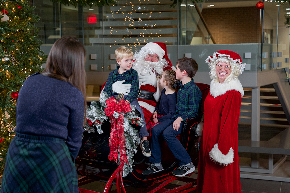 Three children sitting in a sleigh with Santa while their mother and Mrs. Claus watch.