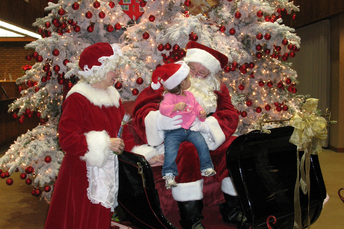 Santa and Mrs. Claus sitting in a sleigh visiting with a little girl in a Santa hat.