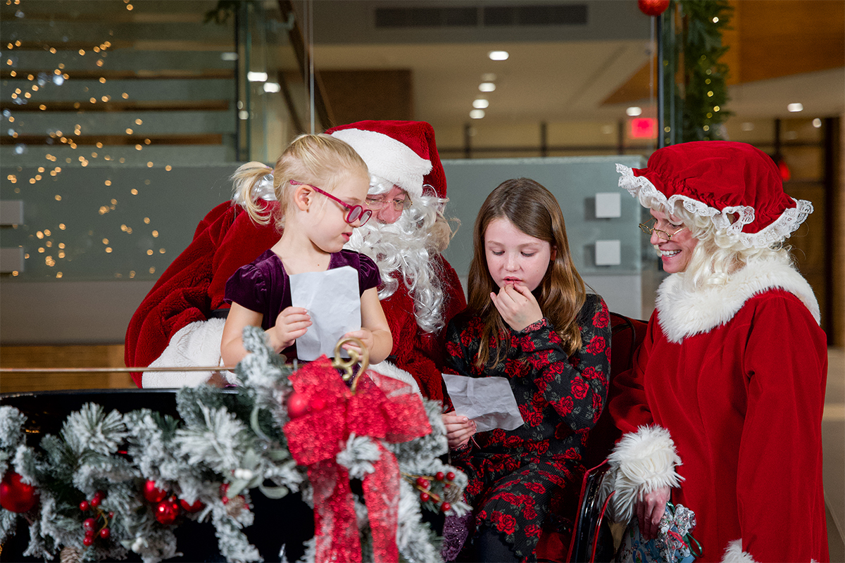 Two young girls read their Christmas wishlists to Santa and Mrs. Claus while they sit in his sleigh.