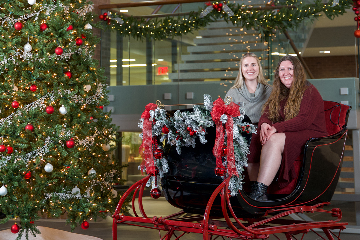 Two women sitting in Santa's sleigh next to the First National Bank Christmas tree.